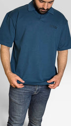 A man wearing a dark teal blue short-sleeve polo shirt with a small embroidered logo on the left chest, paired with dark blue jeans, shown against a light gray studio background.