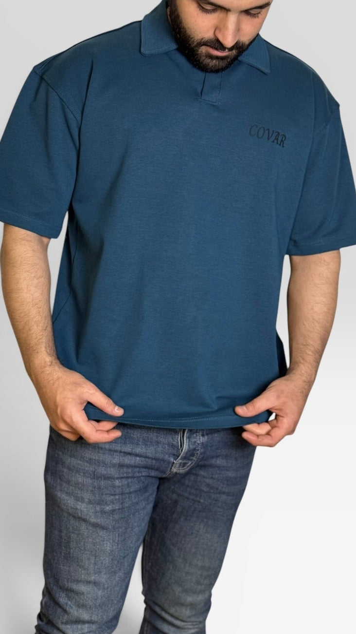 A man wearing a dark teal blue short-sleeve polo shirt with a small embroidered logo on the left chest, paired with dark blue jeans, shown against a light gray studio background.